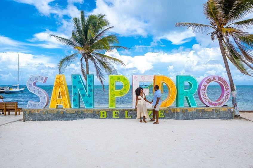 Een strand met een bord waarop 'SAN PEDRO BELIZE' staat en palmbomen. 1