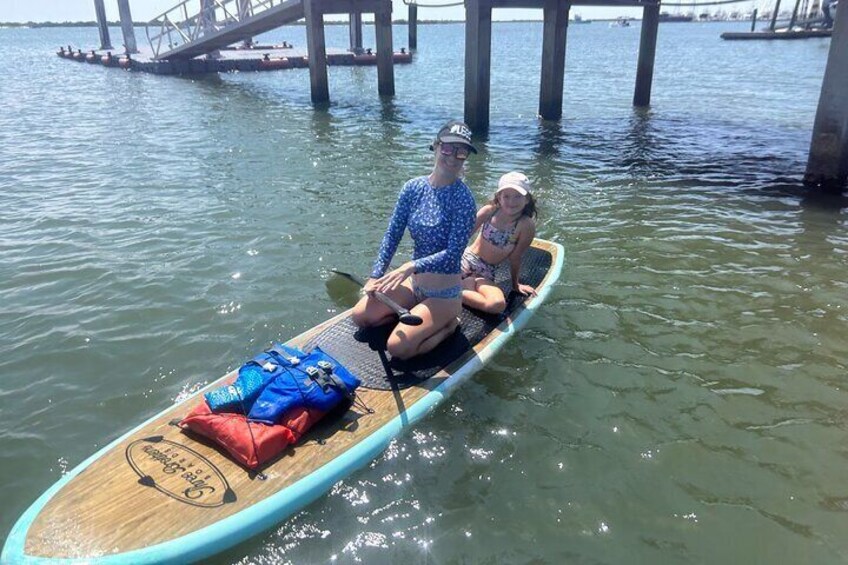 Two people sitting on a paddleboard in a body of water with a bridge in the background. 2