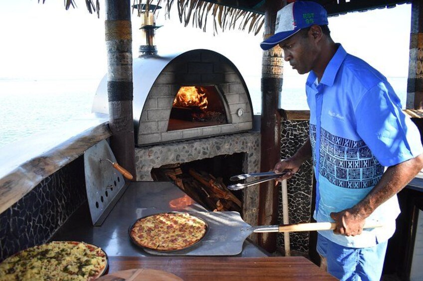 A man operating a brick oven pizza oven, with pizzas being prepared. 3