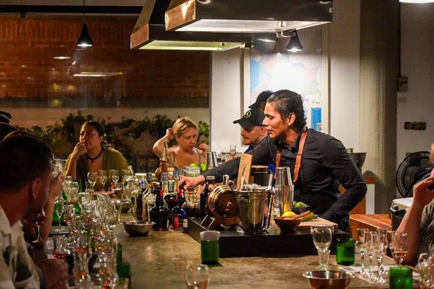 En bar med folk som sitter vid borden, en bartender som blandar drinkar och olika flaskor och glas på bardisken. 2