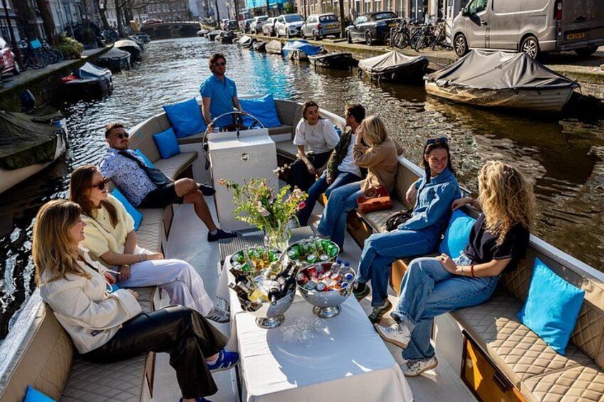 A group of people on a boat with a table of drinks and snacks, surrounded by a canal with other boats and buildings. 1