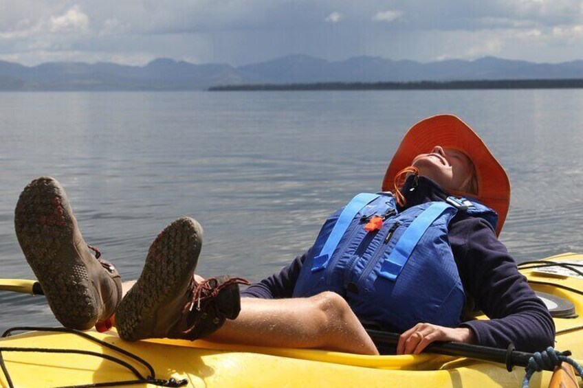 A person lying on a yellow kayak with a blue life jacket and orange hat, wearing brown shoes, on a calm body of water with mountains in the background. 1