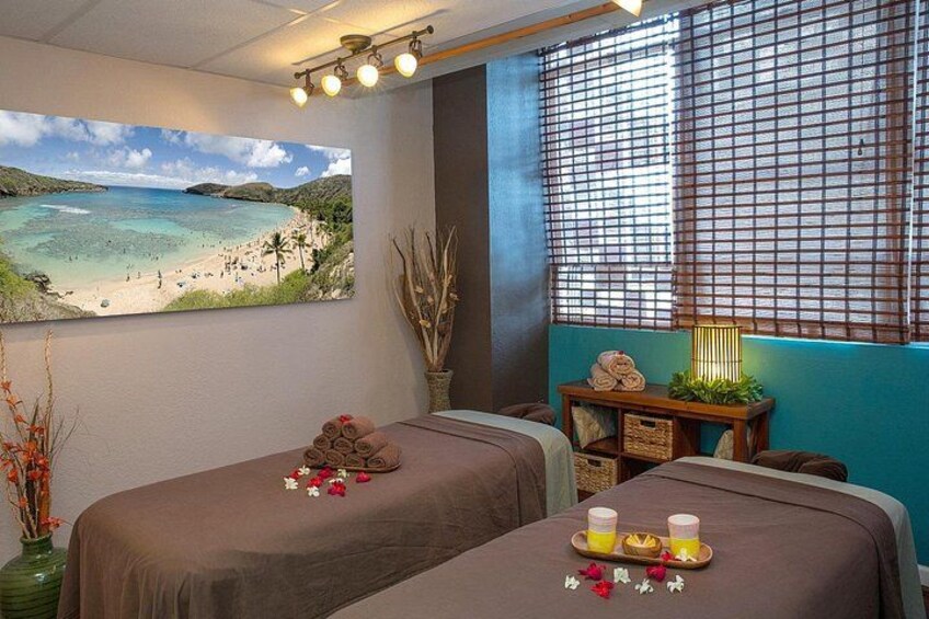 A spa room with two massage tables, a beach painting, a window with blinds, a vase with dried plants, and a tray with towels and drinks. 5