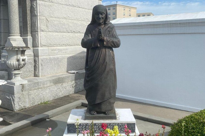 Eine Statue einer Person in einer Robe mit einem Kreuz, stehend auf einem Sockel mit Inschriften und Blumen. 4