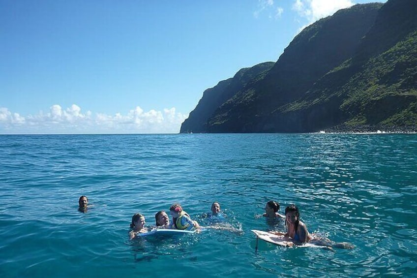 People swimming and surfing in clear blue water with a mountainous coastline in the background. 1