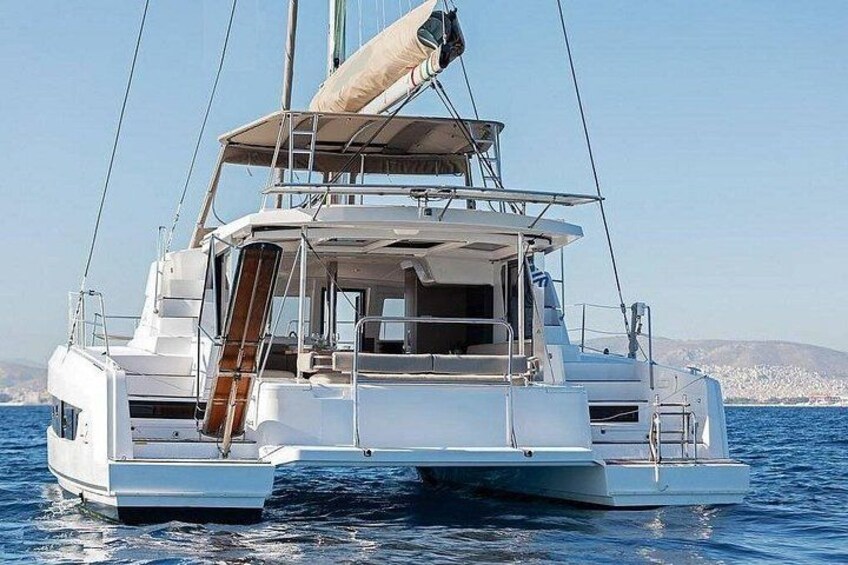 A white catamaran with a cabin and deck seating, sailing on calm waters with a coastline in the background. 2