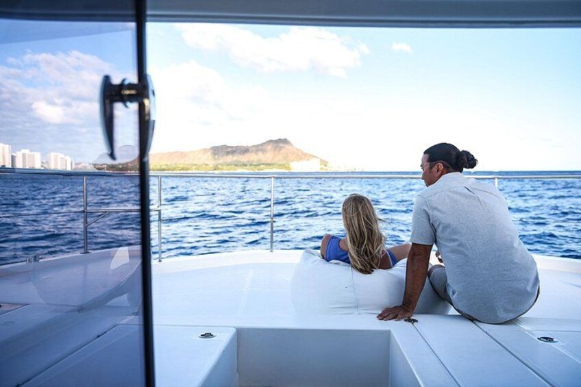 Ein Boot mit zwei Personen, die auf dem Deck sitzen und auf ein Gewässer mit einem entfernten Landteil und Gebäuden blicken. 4