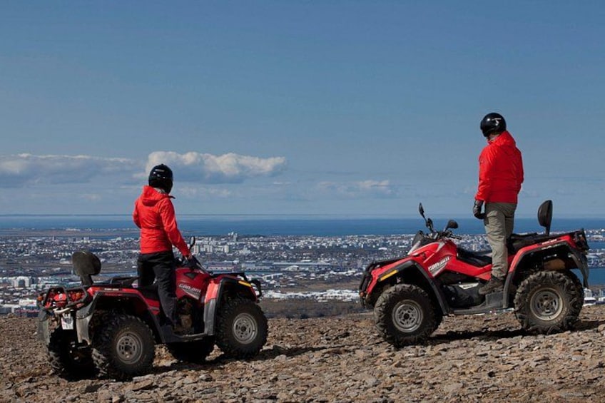 Two people on ATVs on a rocky terrain with a cityscape in the background. 3