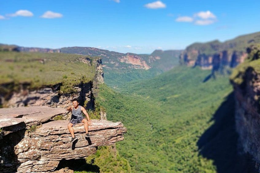 A person sitting on a rock ledge overlooking a canyon with steep cliffs and lush greenery. 3
