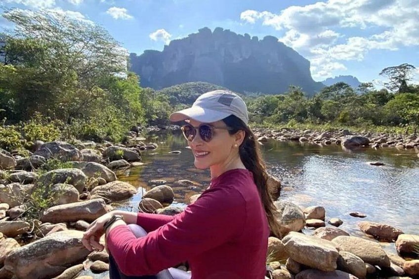 A person sitting by a river with rocks, trees, and a mountain in the background. 1