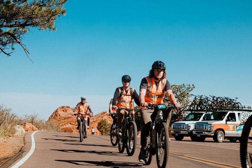 Three cyclists on a paved road with a vehicle carrying bicycles in the background. 5
