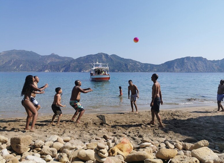 Un gruppo di persone gioca a pallavolo sulla spiaggia, con una barca sullo sfondo e montagne all'orizzonte. 3