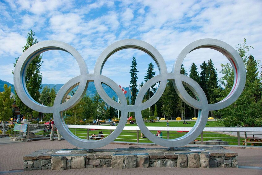 En stor skulptur i form av de olympiska ringarna, placerad i en park med träd och berg i bakgrunden. 2