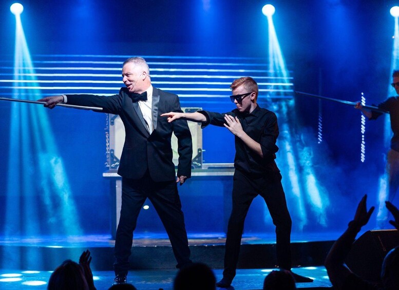 Two men in black suits performing a balancing act on a stage with a blue-lit background. 3
