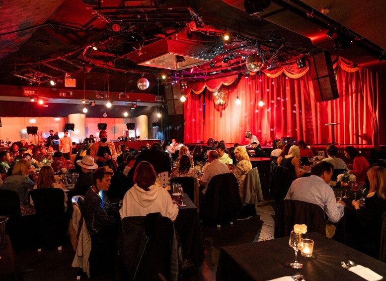A crowded indoor event with tables and chairs, a stage with curtains, and overhead lighting. 1
