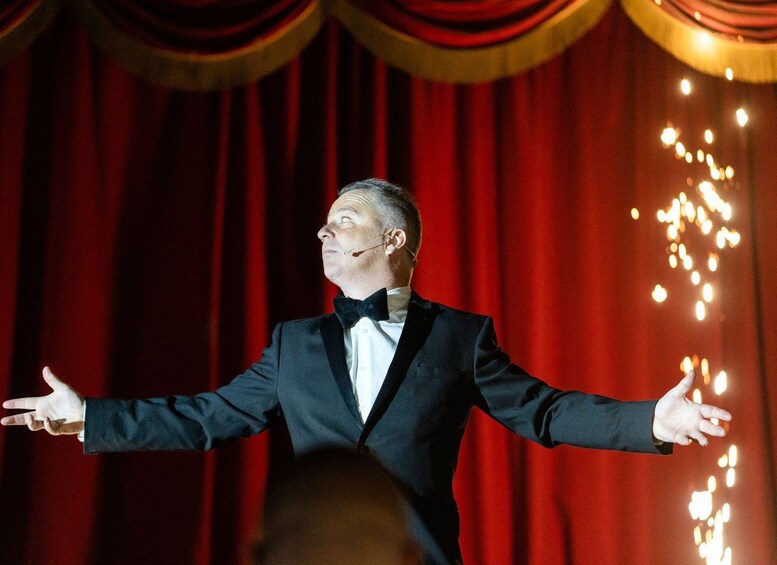 A person in a black suit with a bow tie, standing with arms outstretched in front of a red curtain backdrop. 5