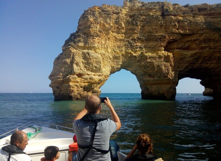 A boat with people taking a photo of a large rock formation with a natural arch over the water. 1
