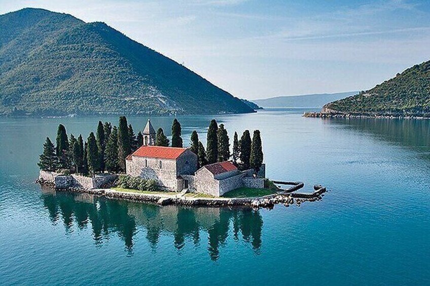 A small island with a church and a red-roofed building surrounded by trees, located in a calm lake with mountains in the background. 3