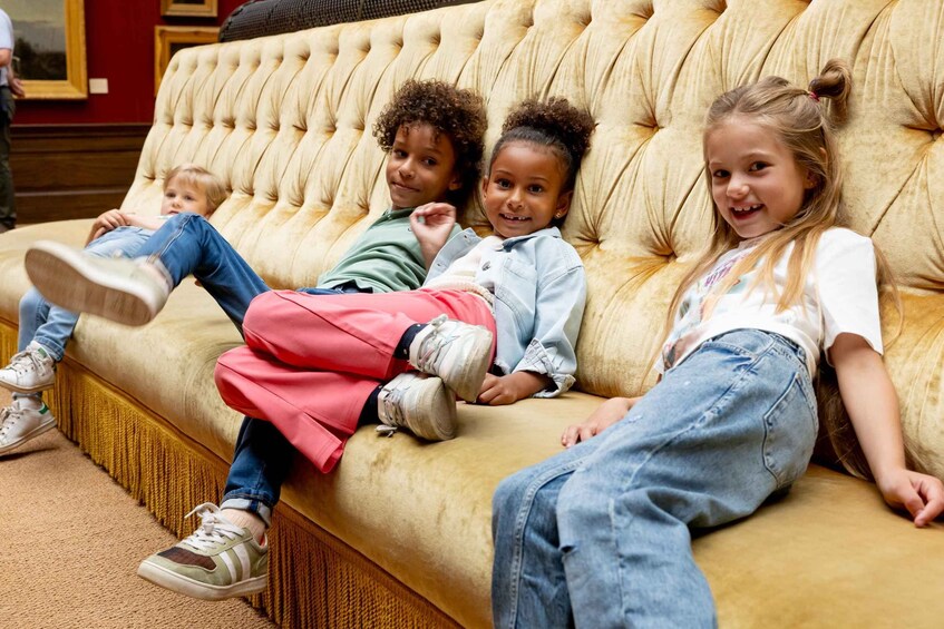Four children sitting on a tufted sofa in a room with a red wall and a framed picture. 2