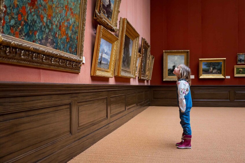A child standing in a hallway with multiple framed paintings on the wall. 5