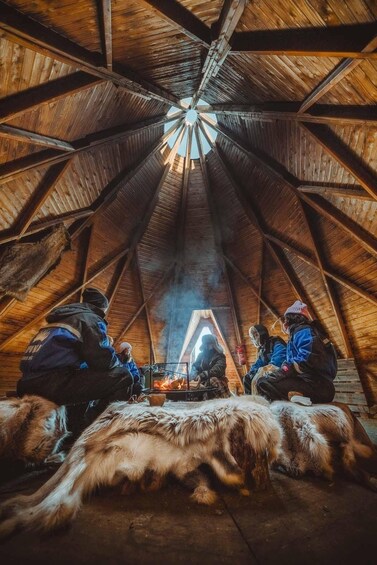 An interior of a wooden structure with a high, star-shaped ceiling and a fire in the center. 3