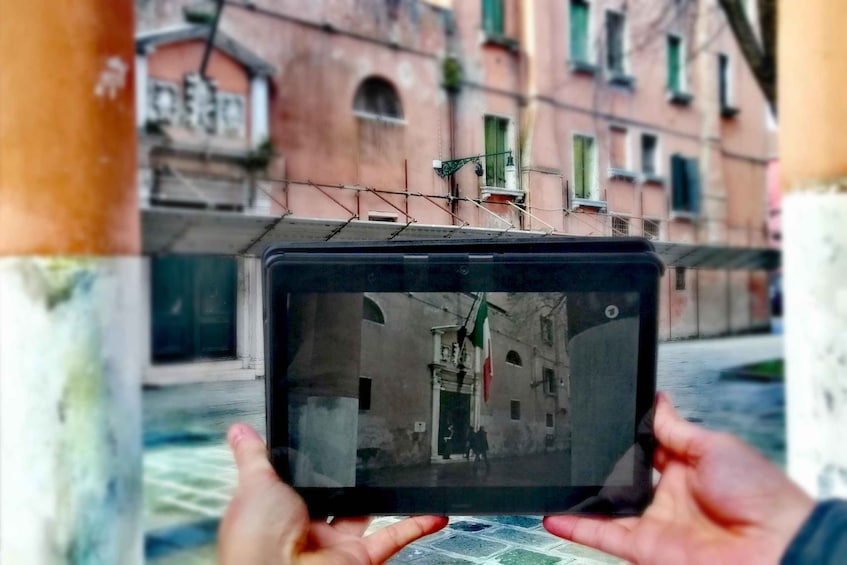 Eine Person hält ein Tablet, das eine Straßenszene mit Gebäuden und einer Flagge zeigt. 1
