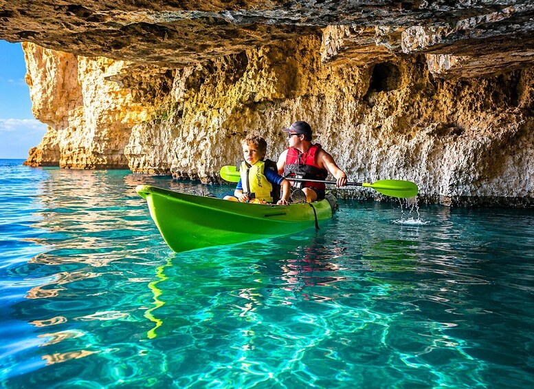 Two people kayaking in a green boat with clear blue water and rocky cliffs in the background. 1