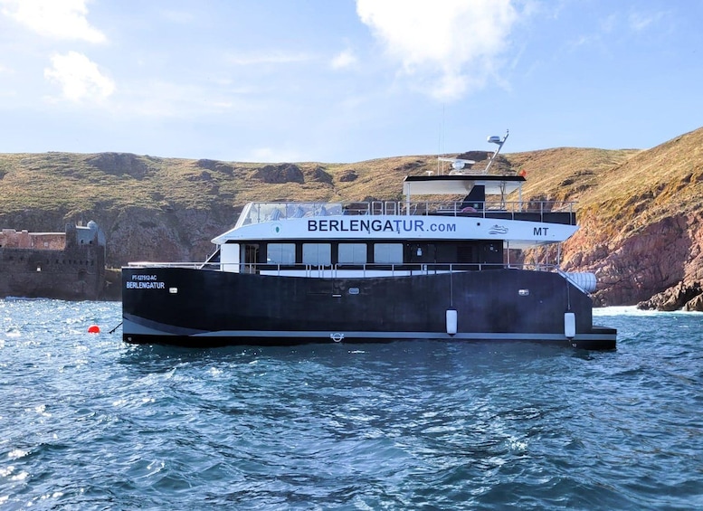 A boat named BERLENGATUR.COM on calm waters with a rocky coastline and a castle in the background. 1