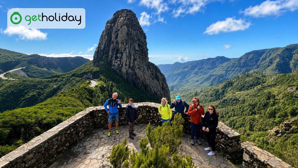 A group of people standing on a stone platform overlooking a mountainous landscape with a prominent rock formation. 1
