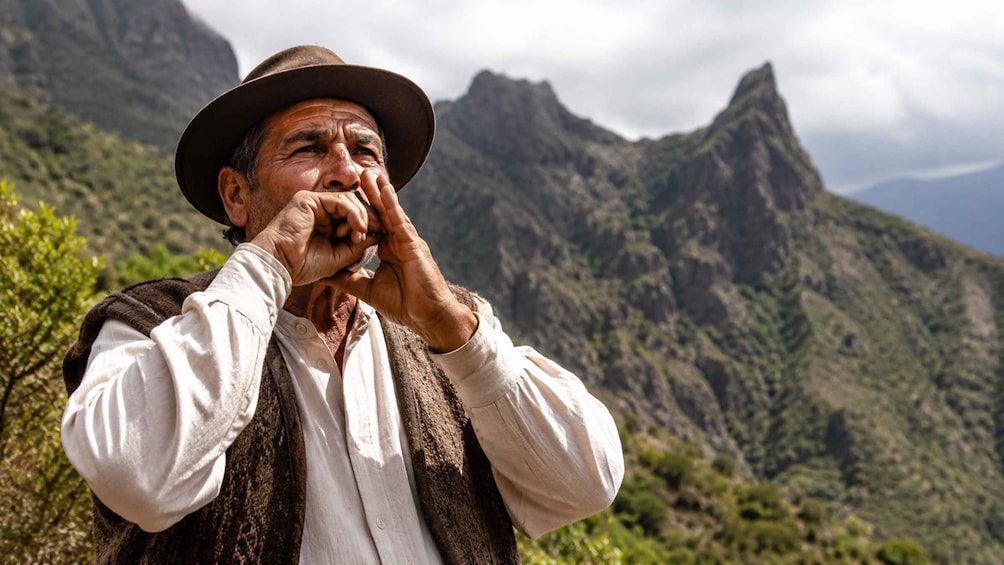 A person in a hat and vest standing outdoors with a mountainous landscape in the background. 2