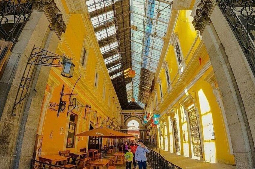A narrow indoor corridor with yellow walls, a glass ceiling, and wooden tables. 4