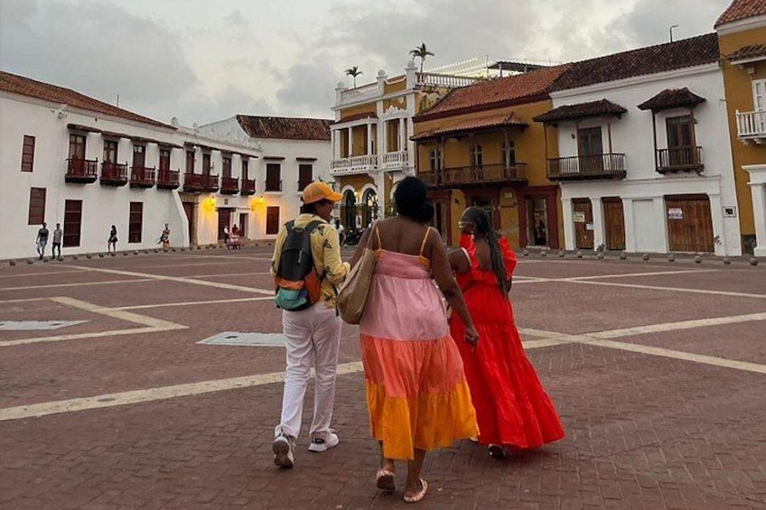 Una plaza con edificios coloniales, una persona con una mochila y mujeres con vestidos coloridos. 4