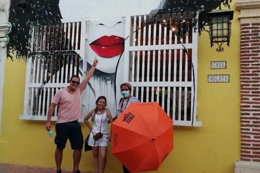 Un mural que muestra el rostro de una mujer de labios rojos, un hombre y una mujer de pie frente a él, y un edificio amarillo con un letrero que dice 'CASA MULATA'. 2