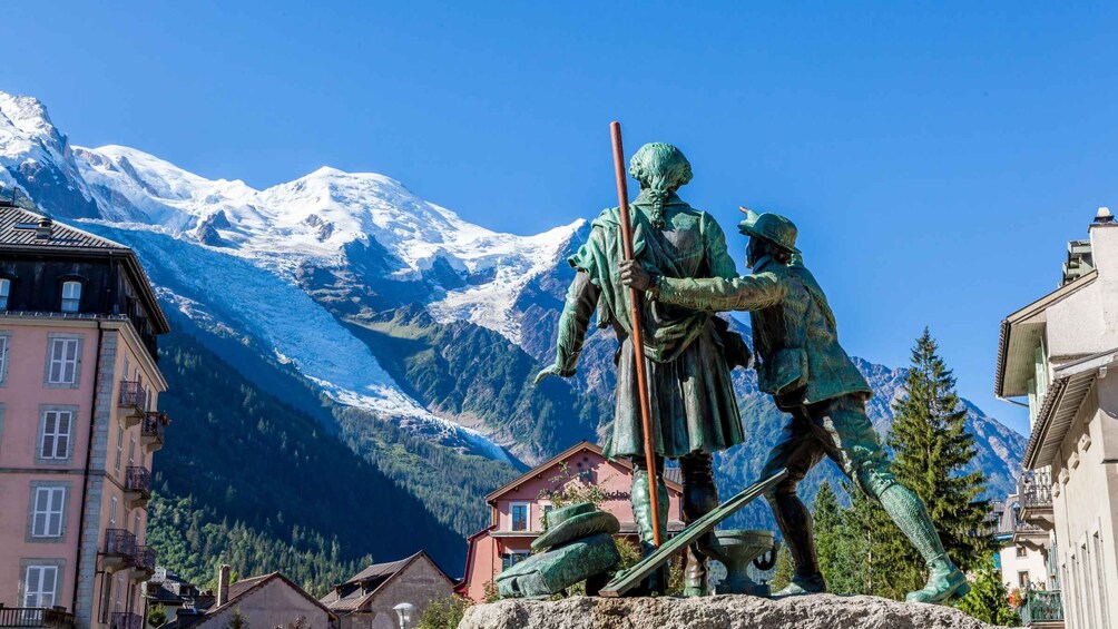 A statue of two figures in historical attire, a mountain range with snow-capped peaks, and traditional buildings in a valley. 2