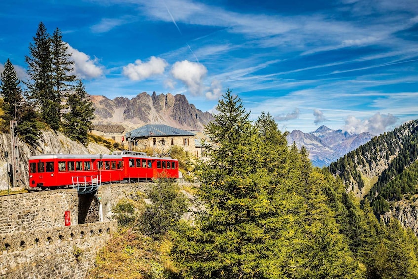 A red train on a mountain track, surrounded by trees and rocky mountains. 1