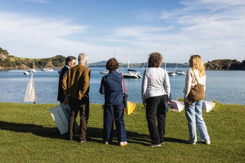 A group of people standing on grass by a lake with boats in the background. 4
