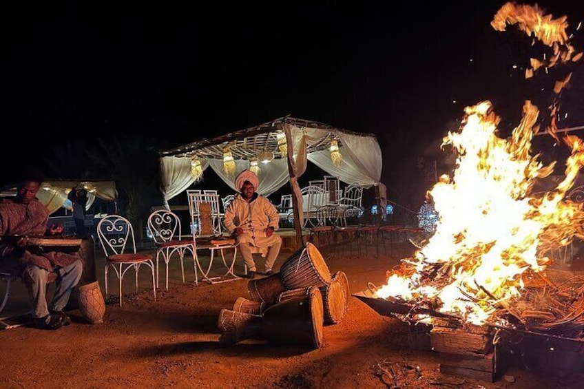 A person sitting by a campfire at night with a tent and chairs in the background. 3