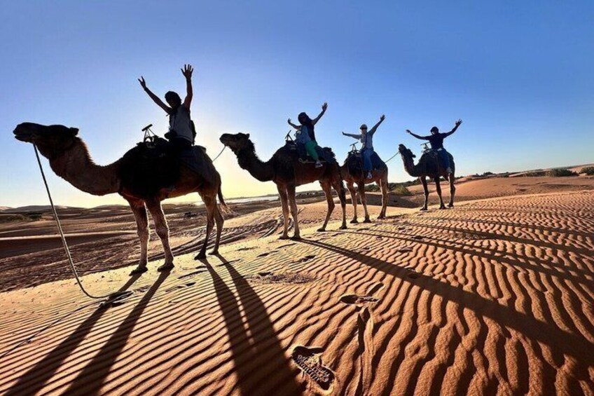 Four people riding camels in a desert with sand dunes. 4