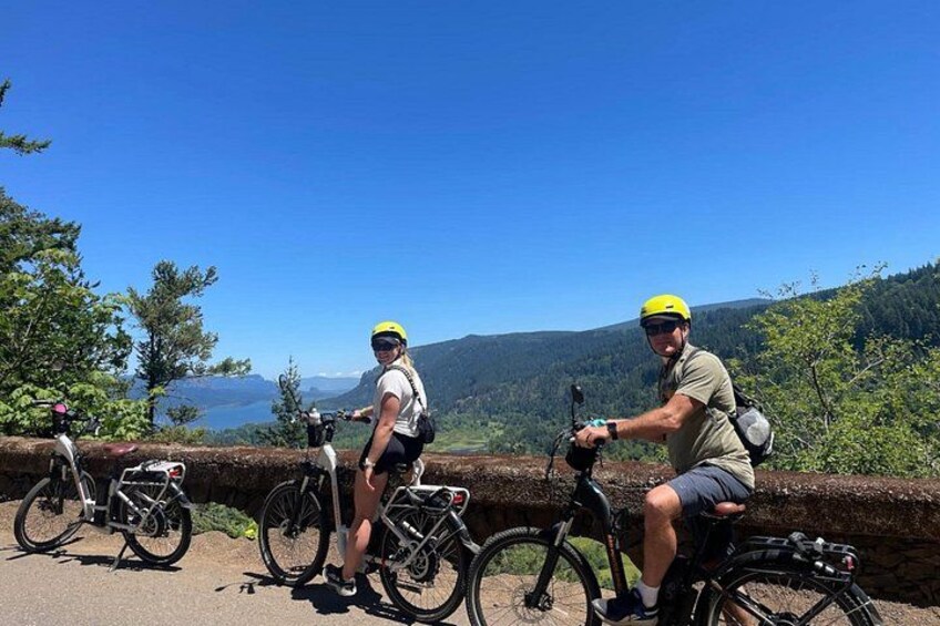 Two cyclists on a paved path with bicycles parked on the side, overlooking a mountainous landscape with a clear sky. 4