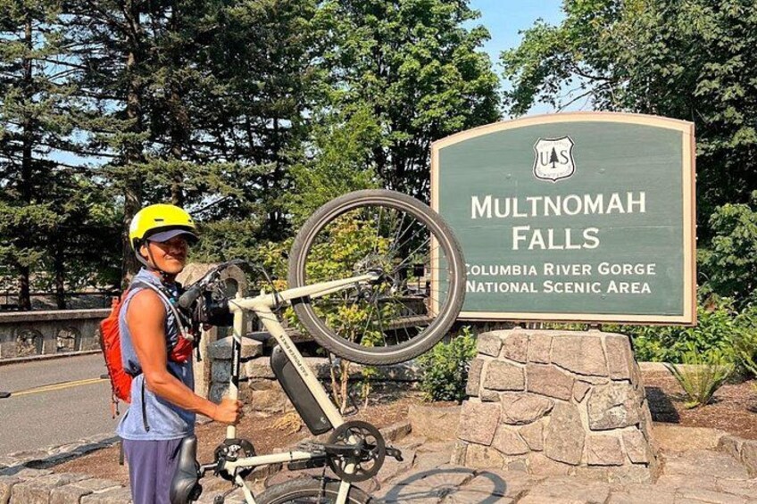 A person with a bicycle and a helmet near a sign for Multnomah Falls. 2