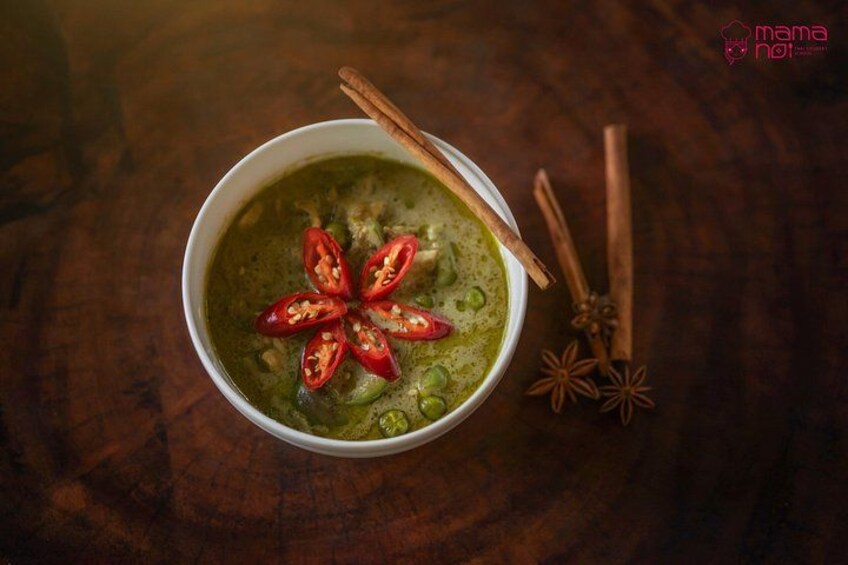 A bowl of green soup garnished with chili slices and cinnamon sticks on a wooden surface. 2