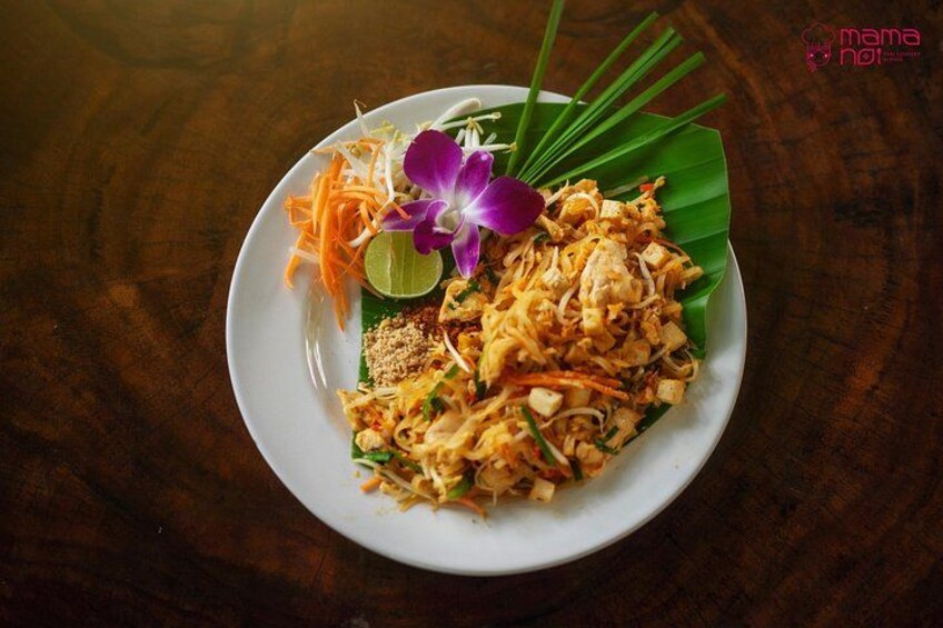 A plate of Pad Thai with a lime wedge, carrot garnish, and a purple flower on a banana leaf. 4