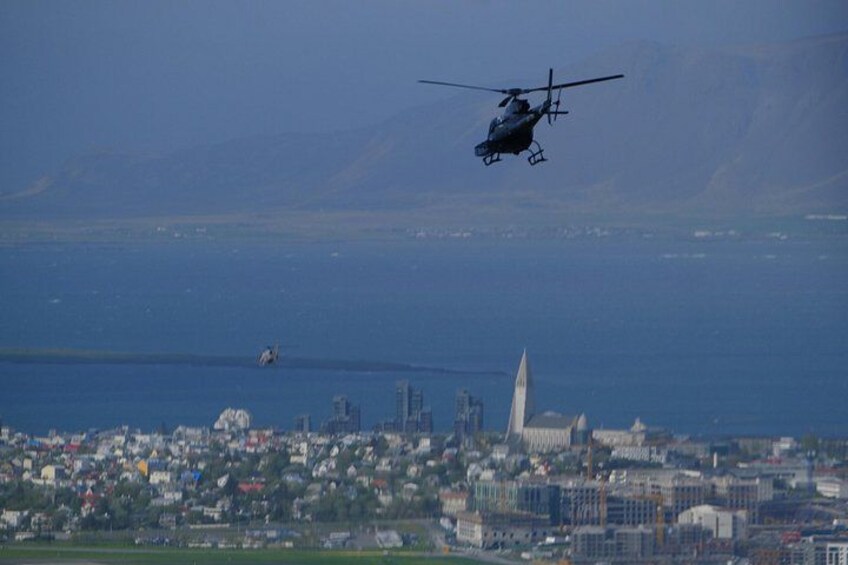 Een helikopter vliegt boven een kustplaats met gebouwen en een opvallende toren. 5