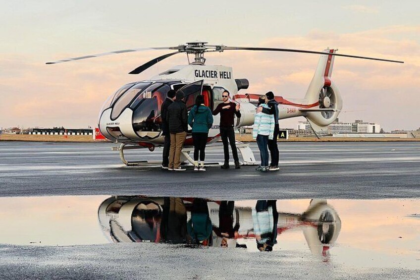 Ein mit dem Schriftzug 'GLACIER HELI' versehener Hubschrauber steht auf einer feuchten Fläche, umgeben von Personen. 2
