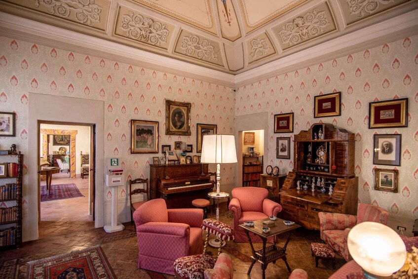 A vintage living room with patterned wallpaper, a piano, armchairs, and a bookshelf. 5