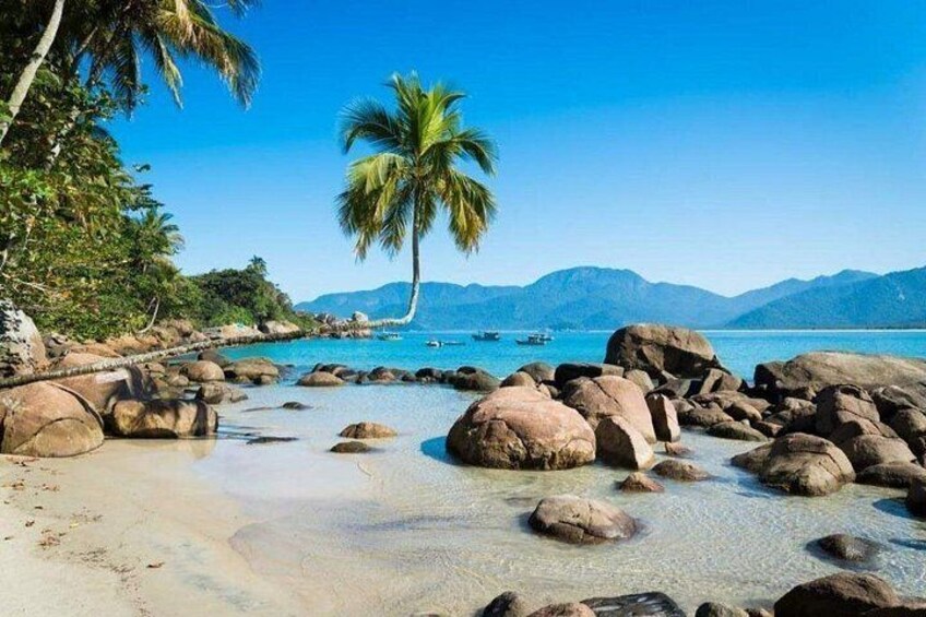 A tropical beach with large rocks, clear water, palm trees, and distant mountains. 5