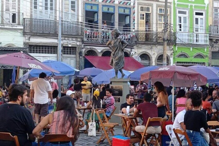 A crowded outdoor market with people sitting at tables under umbrellas, a statue in the center, and colorful buildings in the background. 3