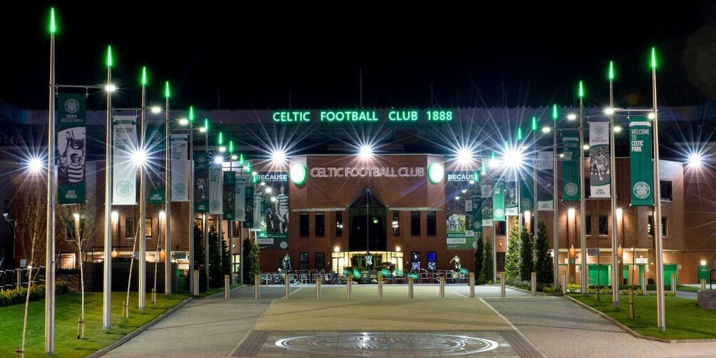 The entrance to Celtic Football Club stadium at night, with illuminated green lights and banners. 1