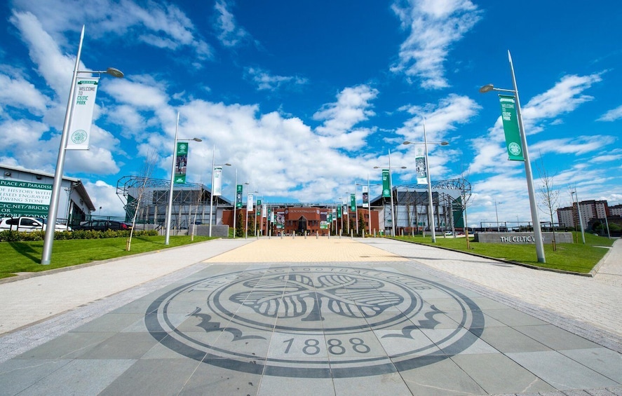 A stadium entrance with a Celtic Park sign, a large circular emblem on the ground, and a clear blue sky. 1