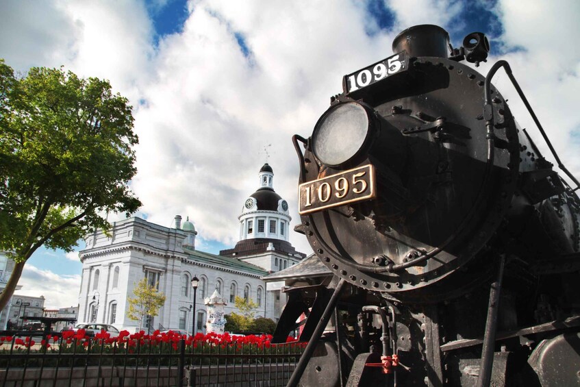 Een stoomlocomotief met het nummer 1095, een historisch gebouw met een klokkentoren, en rode bloemen op de voorgrond. 1